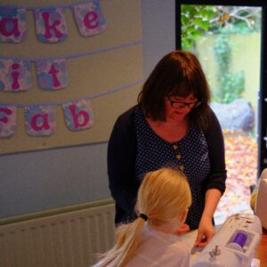 Photo of Abigail (a woman with brown hair and glasses wearing a blue dress with white spots and yellow buttons) giving instruction to child with long blond hair.