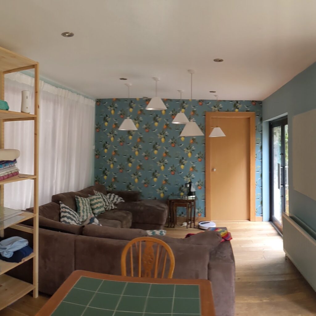 Seating area at Make it Fab. A large brown corner sofa with blue, green and cream scatter cushions. Blue wallpaper on the wall with oranges and lemons